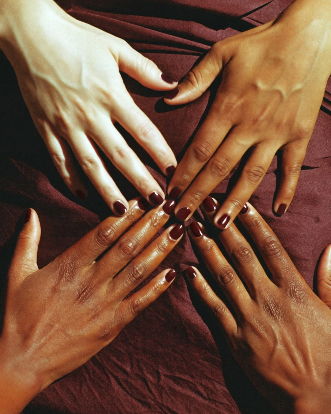 Four hands with oxblood nails meeting at the centre on burgundy silk