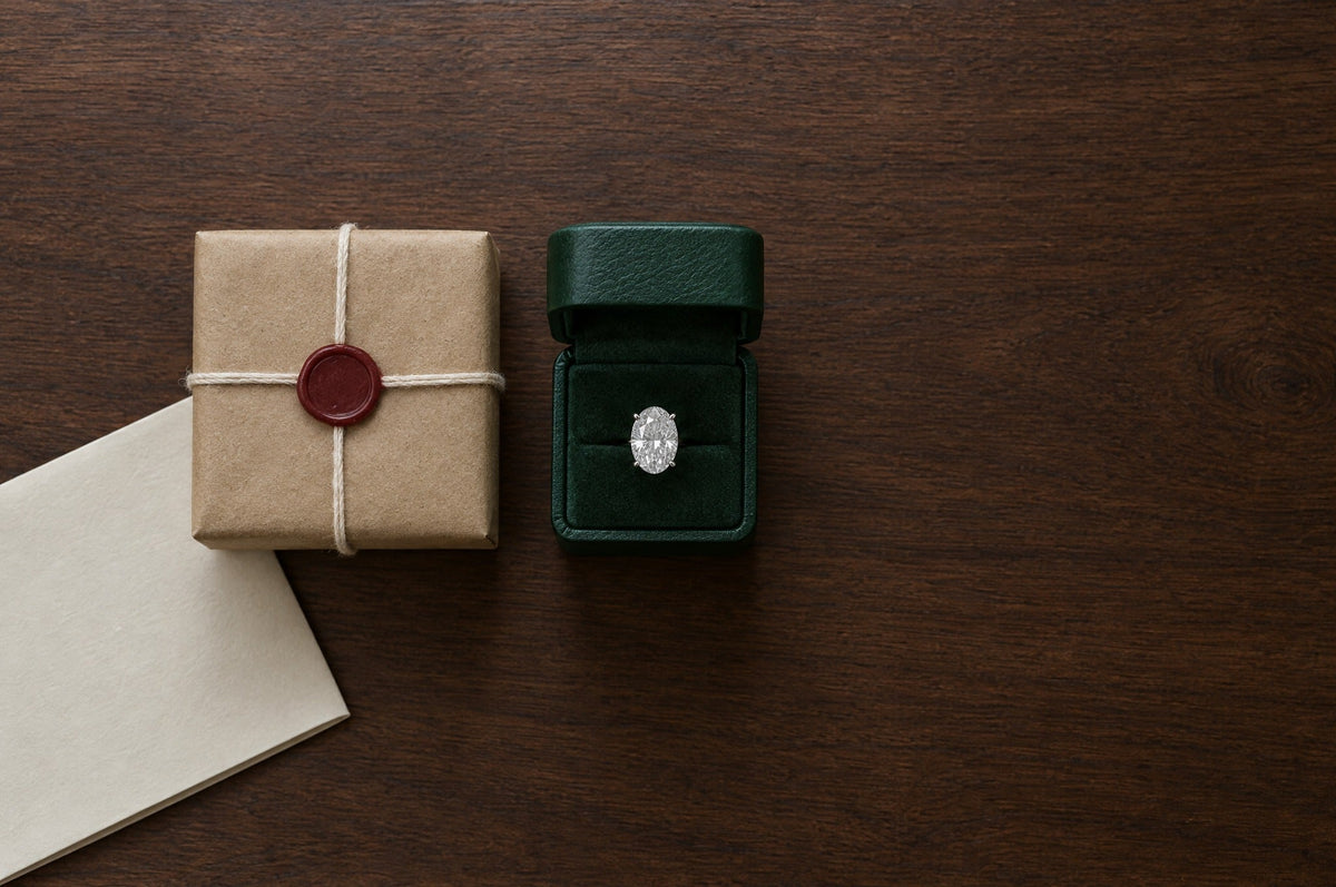 A sealed kraft-paper parcel tied with string beside an open green ring box holding an oval diamond solitaire, seen from above on a walnut desk
